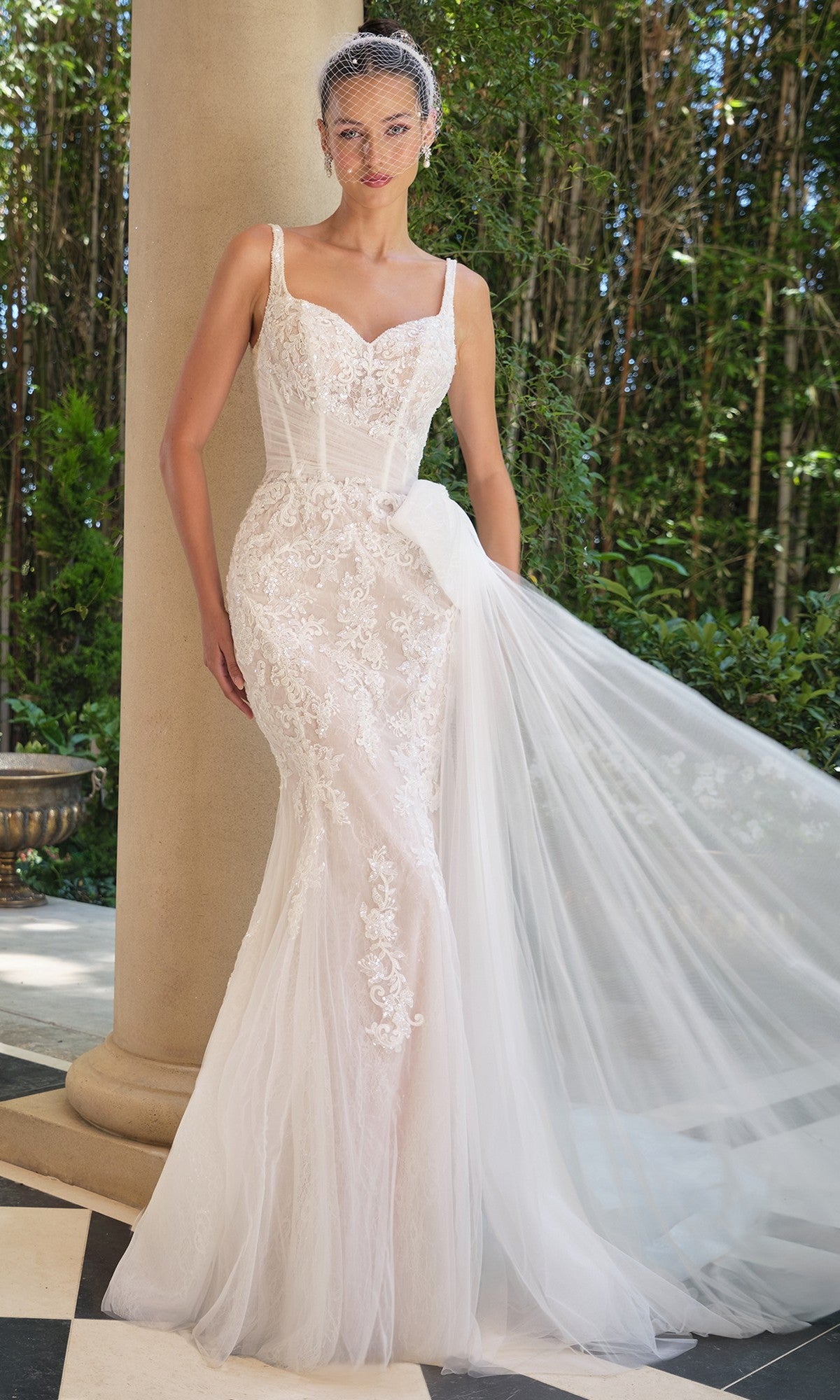A woman stands outdoors in the Andrea and Leo WL068 long wedding dress with a sheer corset bodice, holding a flowing tulle train and wearing a birdcage veil.
